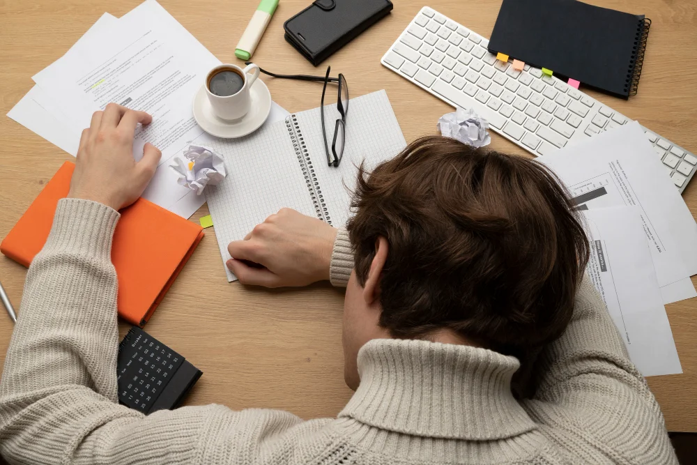 Estudante com a cabeça apoiada sobre a mesa rodeado de cadernos e papéis amassados, representando a procrastinação nos estudos por esgotamento e falta de progresso visível.