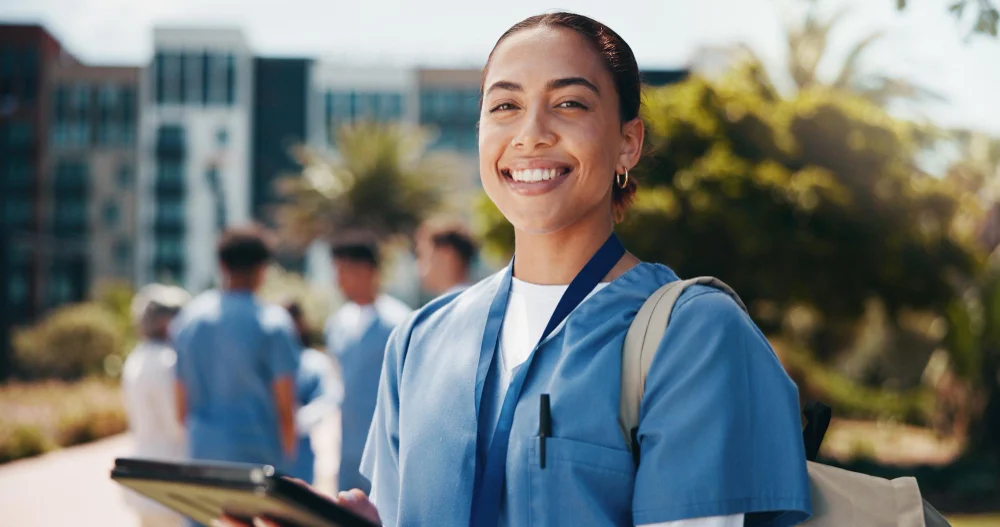 Estudante de Medicina em federal sorrindo, de pijama cirúrgico azul e tablet na mão, no campus da universidade